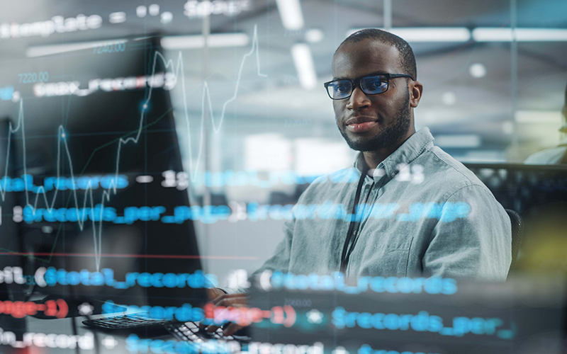 Imagem mostra um homem negro, utilizando um computador. Por sobre a foto, há ícones e imagens que nos remete ao ambiente tecnológico e de códigos de programação