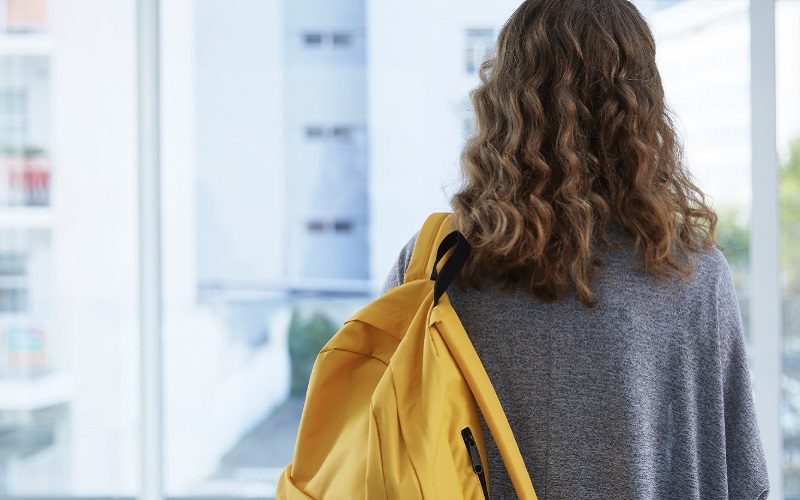 Aluna chegando na escola, de costas, de mochila amarela.