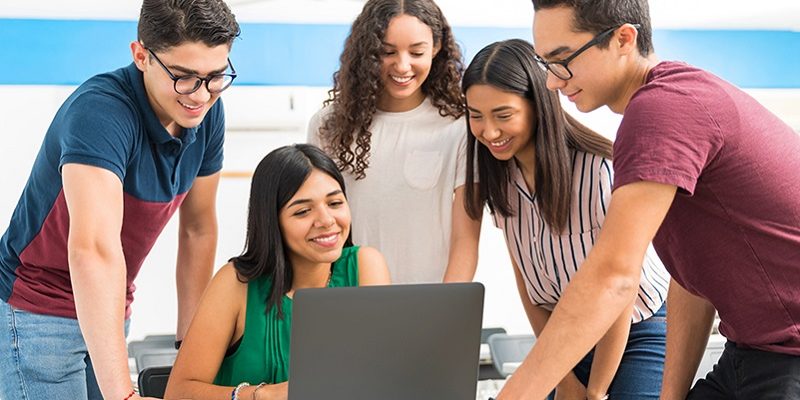 Imagem mostra uma professora usando um notebook, enquanto quatro jovens estudantes estão ao seu lado acompanhando o que ela está fazendo. Todos são brancos e estão sorrindo
