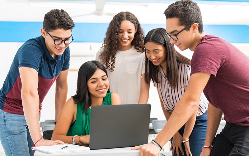 Imagem mostra uma professora usando um notebook, enquanto quatro jovens estudantes estão ao seu lado acompanhando o que ela está fazendo. Todos são brancos e estão sorrindo