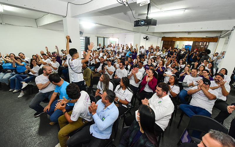 Imagem mostra grupo de voluntários e de estudantes reunidos em um espaço fechado durante o Dia dos Voluntários. As pessoas estão aplaudindo a fala de um jovem que está em pé à frente de todos