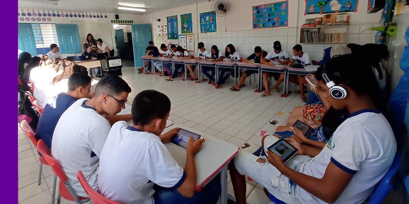 Imagem mostra um grupo de estudantes em sala de aula utilizando tablets e fones de ouvido