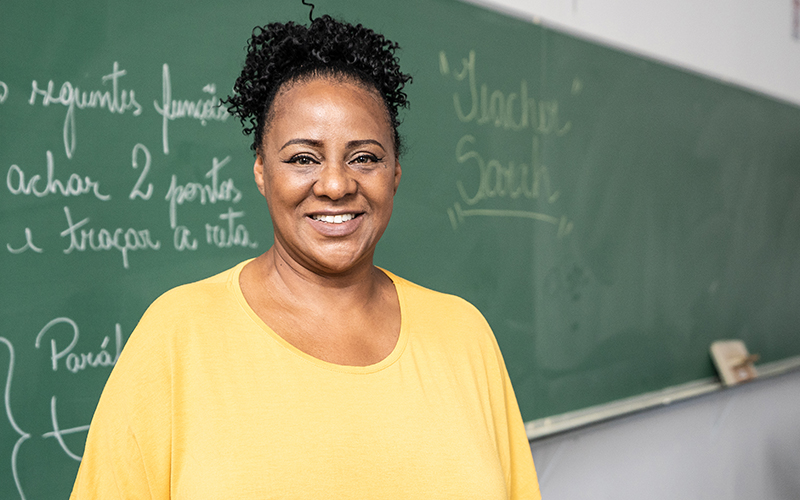 Imagem mostra uma professora negra em sala de aula, sorrindo para a foto. Ela usa blusa amarela e cabelos presos. Ao fundo, há uma lousa