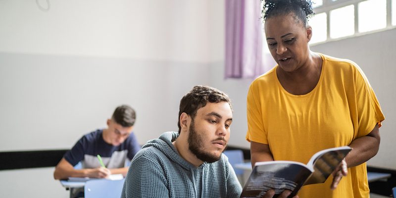 Em sala de aula, professora ajuda individualmente aluno, com um livro na mão.