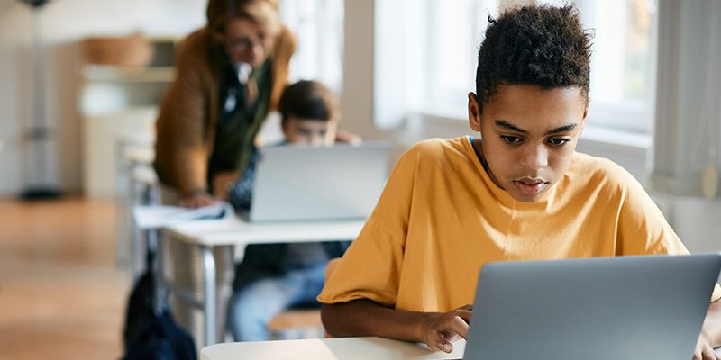 Dois estudantes diante de computadores na sala de aula, com professora em pé ao fundo