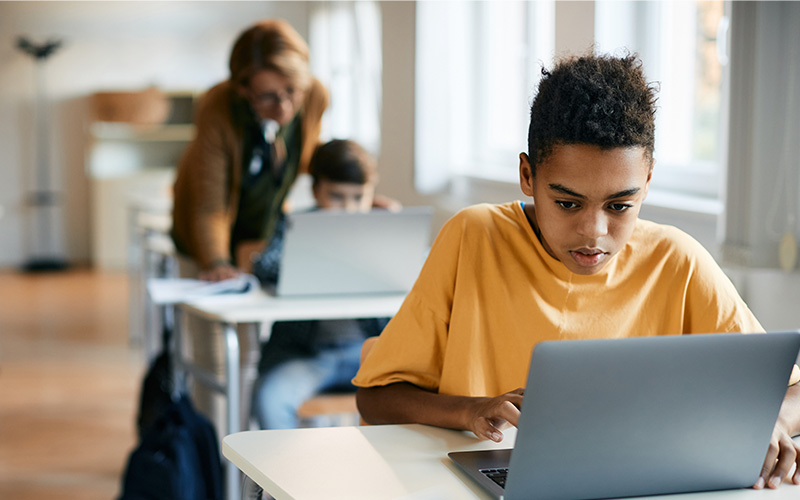 Dois estudantes diante de computadores na sala de aula, com professora em pé ao fundo