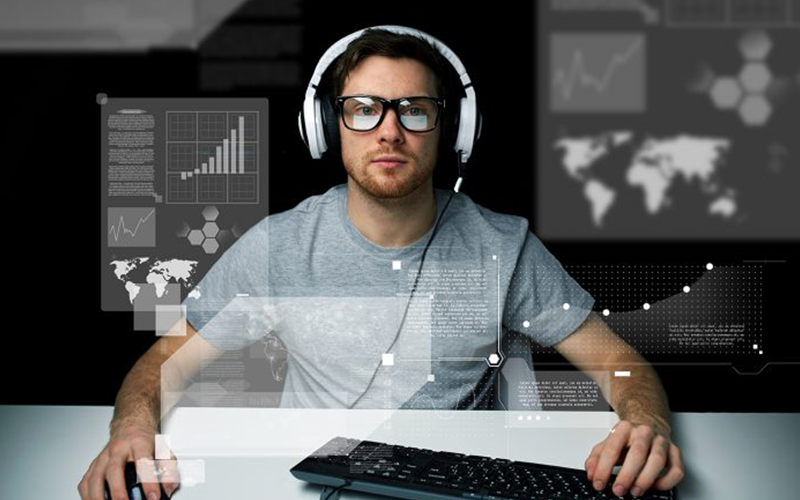Jovem branco, de óculos, com fones de ouvido brancos utiliza um computador