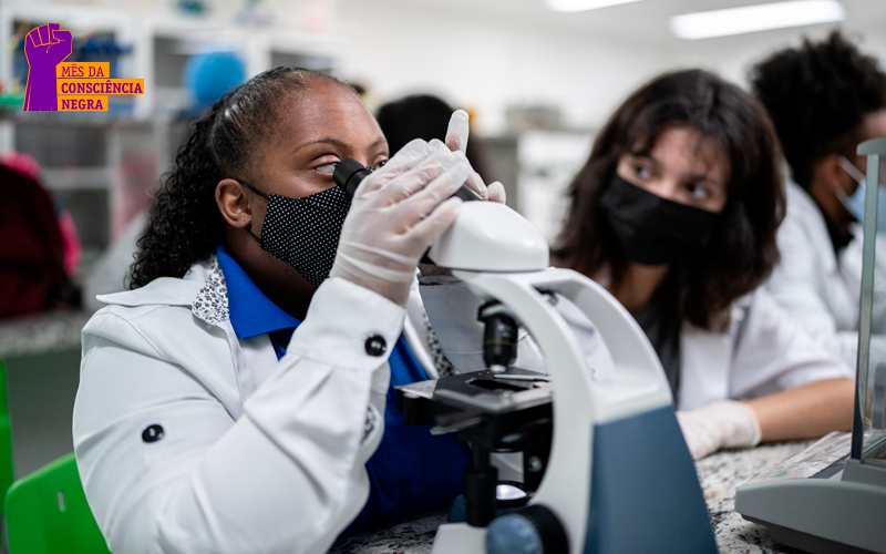 Cientista negra, vestida com jaleco branco, olha em um visor de mi-croscópio.