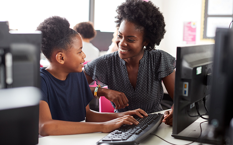 Imagem mostra uma professora negra ao lado de uma aluna negra. Elas estão utilizando um computador