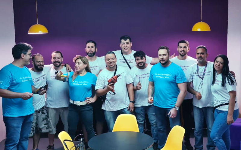 Imagem mostra um grupo de 12 voluntários; são 10 homens e 2 mulheres. Eles estão reunidos em uma sala, ao fundo há uma parede na cor roxa. Eles usam camisetas com a frase sou voluntário telefônica