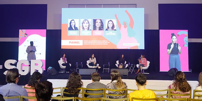 Imagem mostra o palco onde aconteceu o festival social good brasil, com quatro palestrantes sentadas. Há telões e também é possível ver a plateia