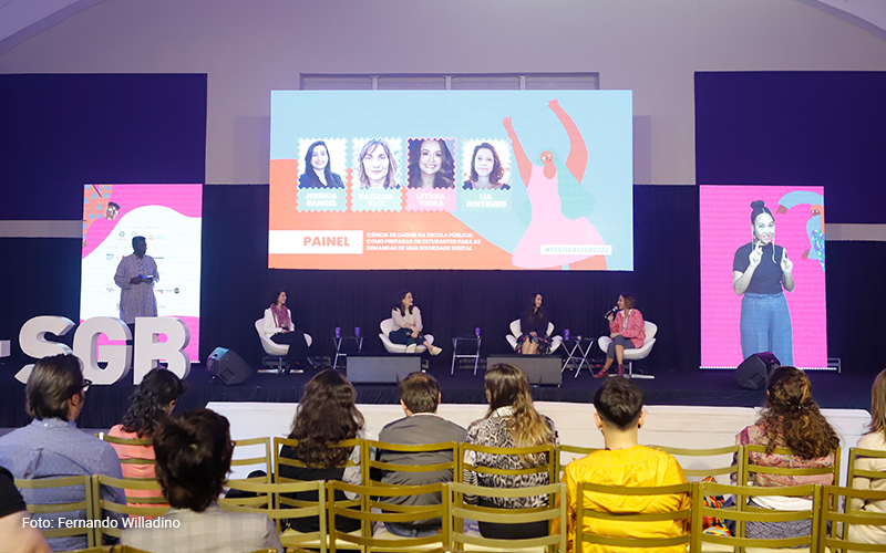 Imagem mostra o palco onde aconteceu o festival social good brasil, com quatro palestrantes sentadas. Há telões e também é possível ver a plateia