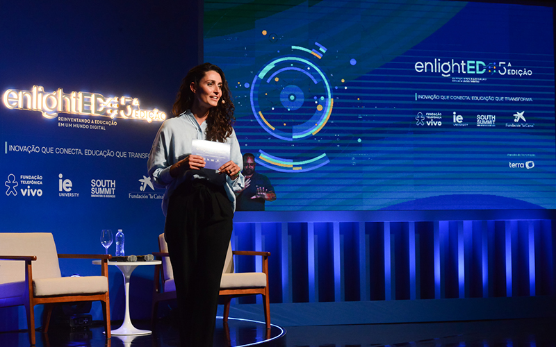 Imagem mostra a apresentadora do evento no palco. Ela é branca, usa um microfone e está falando á plateia. No palco há telões com logo do evento, predominantemente na cor azul