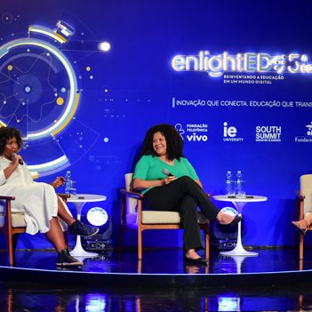 Imagem mostra o palco do enlighted com três palestrantes no palco. Elas são mulheres negras, estão sentadas, uma delas está com o microfone falando ao público