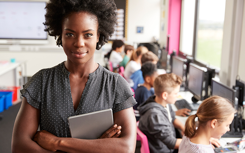 Imagem mostra uma professora negra em primeiro plano, olhando para a câmera. Ela está a frente de um grupo de alunos, que estão usando computadores em sala de aula