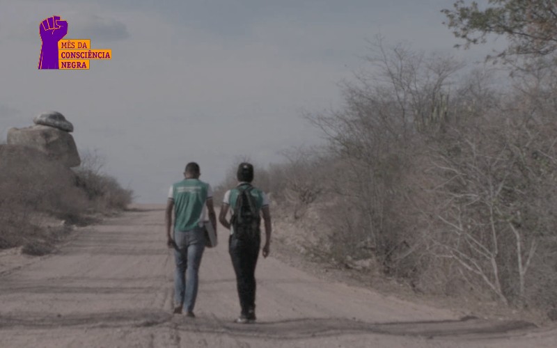 A imagem mostra dois jovens de costas, andando por uma estrada de terra, numa paisagem do semiárido do Ceará. O rapaz da esquerda veste calça jeans e camiseta do uniforme escolar verde e está com um caderno na mão direita. O rapaz da direita veste calça jeans e camiseta verde do uniforme escolar e tem uma mochila preta nas costas