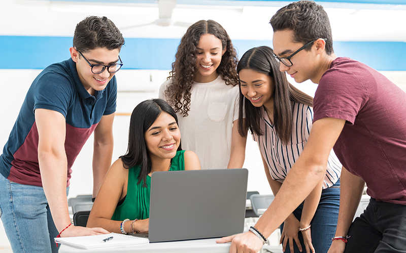 Imagem mostra um grupo de jovens estudantes ao redor de uma professora que está sentada usando um computador. Todos estão em uma sala de aula