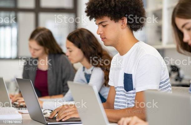 College student using laptop in class during computer lesson. Side view of guy sitting in a row studying in classroom on pc. Black high school student studying on laptop in university library.
