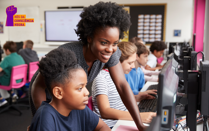 Imagem mostra uma sala de computadores com adolescentes. Em primeiro plano está uma estudante olhando para a tela do computador. Atrás dela, em pé está a professora, sorrindo e mexendo no mouse.
