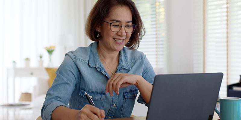 Imagem mostra uma professora branca estudando com o uso de um notebook. Ela segura ma caneta com a mão direita e faz anotações em um caderno, que está sobre a mesa