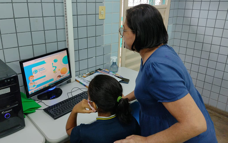 Professora ensina aluna diante de computador em sala de aula