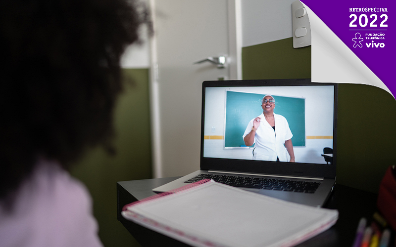 De costas, menina olha para a tela de um computador, onde uma professora dá aula em frente à lousa.