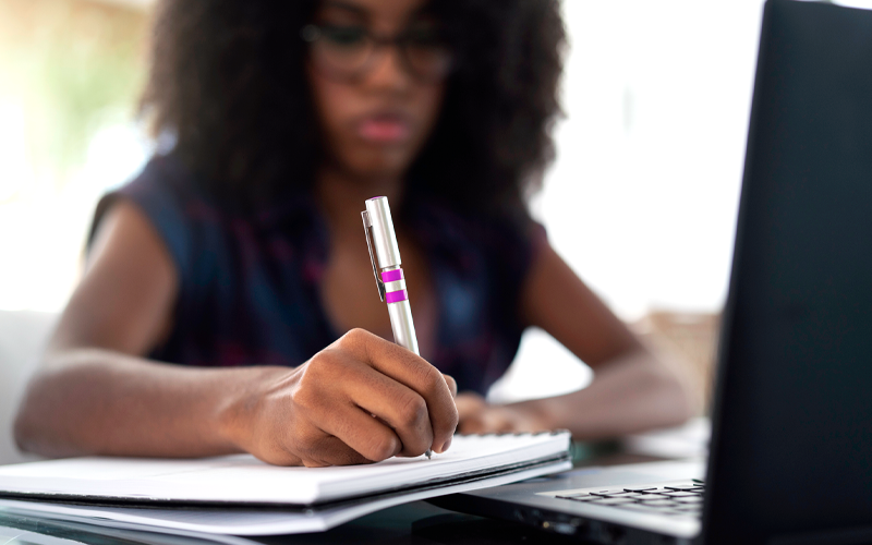 Imagem mostra uma educadora negra escrevendo em um caderno. À sua frente, há um notebook preto