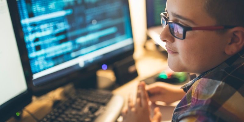 Adolescente usa óculos e camisa xadrez. Está em sentado em frente ao computador, com duas telas.