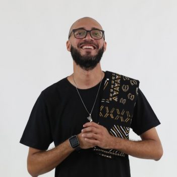 A foto mostra o escritor Israel Neto, fundador da Editora Kitembo. Ele veste camiseta preta, colar e lenço com símbolos e estampas africanas.