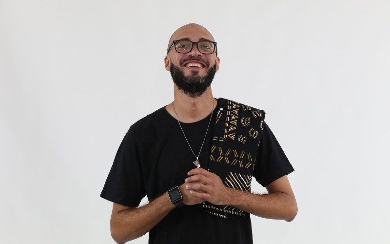 A foto mostra o escritor Israel Neto, fundador da Editora Kitembo. Ele veste camiseta preta, colar e lenço com símbolos e estampas africanas.