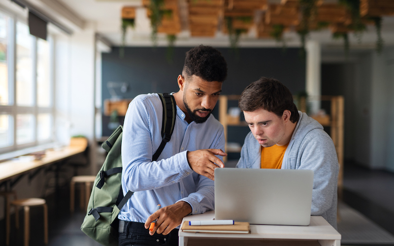 Em uma sala de aula, um estudante negro conversa com um estudante com síndrome de down, em frente a um laptop aberto