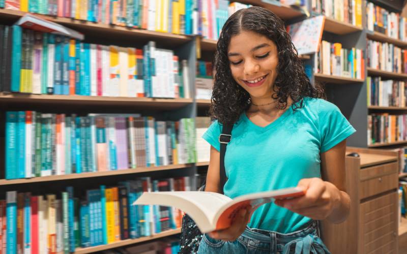 A imagem mostra uma aluna com um livro didático aberto em suas mãos, diante de uma estante de livros