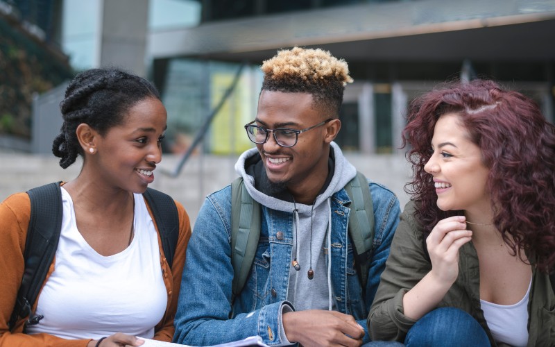 A imagem mostra três jovens de ensino médio. A jovem da direita tem cabelo vermelho, veste blusa de manga comprida verde, camiseta branca e calça jeans e traz a mão levantada na altura do queijo. O jovem do meio usa óculos e tem cabelos pintados de loiro. Está vestindo blusa de moletom cinza, jaqueta jeans e mochila cinza nas costas. A jovem da ponta esquerda tem cabelo preto e com trança, veste camiseta branca, blusa de frio laranja, mochila preta nas costas e está com alguns papeis na mão direita. Os três estão sorrindo.