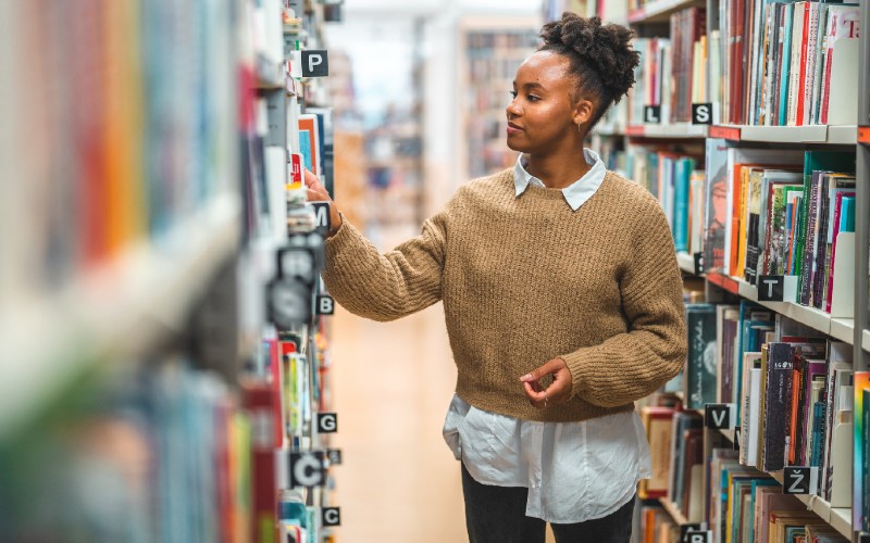 Jovem mulher negra veste camisa azul clara e suéter bege. Ela está na biblioteca e escolhe livros sobre ciência de dados
