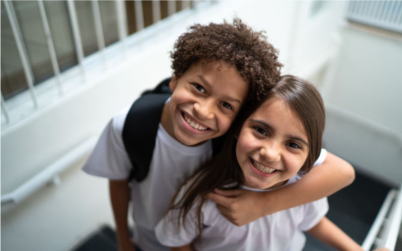 Imagem mostra duas crianças de ensino fundamental 1. O menino está à esquerda da imagem e abraça a menina que está à direita. Os dois usam camiseta branca. O menino está de mochila preta nas costas.