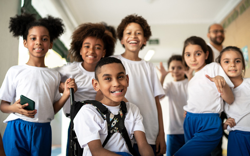 A imagem mostra um grupo de sete crianças da educação infantil. Todos vestem uniforme, com camiseta branca e calça azul royal. Em primeiro plano, está um menino na cadeira de rodas. Os demais estudantes estão sorrindo e em pé em volta dele. Ao fundo do grupo, está a cabeça desfocada de um professor.
