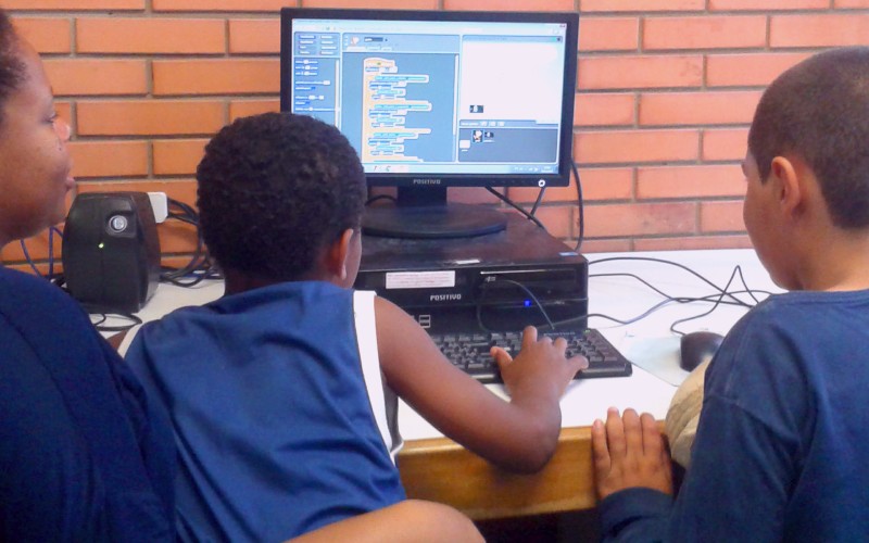 A imagem mostra a professora Nelza Jaqueline Franco com dois de seus alunos do 4º ano da EMEF Afonso Guerreiro Lima, em Porto Alegre. Ela está sentada na ponta da esquerda, o aluno que está no meio está mexendo no teclado e o aluno da ponta direita está perto do mouse. Os três olham atentamente para a tela do computador com o jogo Scratch.