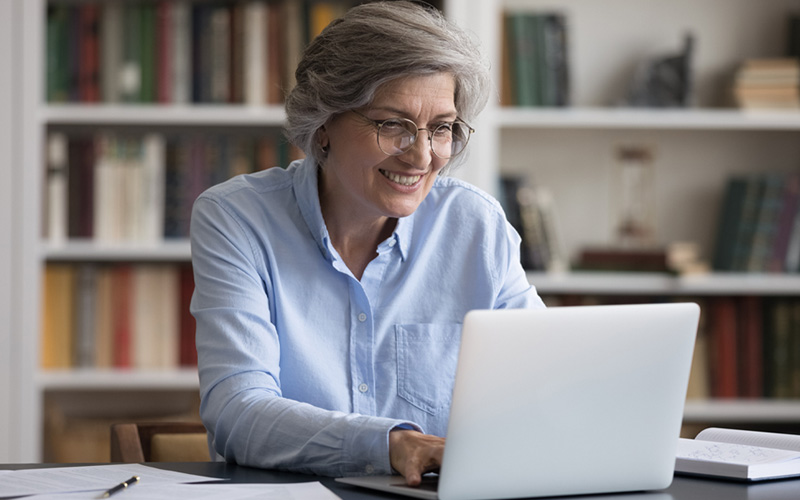 A foto mostra uma mulher sorrindo enquanto olha para um notebook branco. Ela tem cabelos grisalhos e usa óculos de aro redondo. Está vestindo uma camisa de manga longa azul clara. Atrás dela, está uma estante com livros.