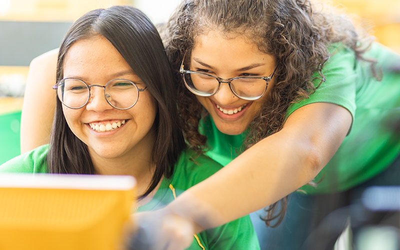 Imagem mostra duas estudantes lado a lado olhando para a tela de um computador. As duas são morenas, usam óculos e camisetas verde
