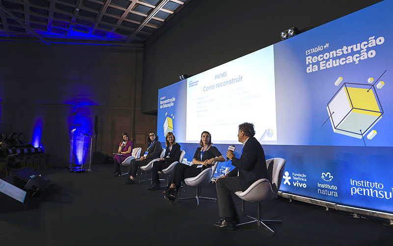 Imagem mostra o palco do evento, com cinco palestrantes sentados em cadeiras sobre o palco