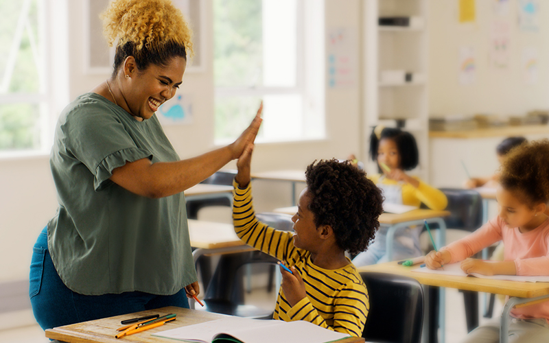 Na imagem, vemos uma professora negra cumprimentando com as mãos um estudante negro, os dois estão sorrindo