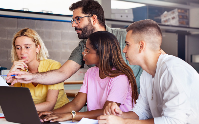 Na imagem, um professor orienta um grupo de três alunos. O professor está em pé e os estudantes estão sentados. Os três olham para a tela de um notebook.