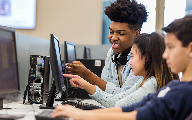 Na imagem, três estudantes estão em sala de aula utilizando computadores. Um dos estudantes é negro e os demais são brancos