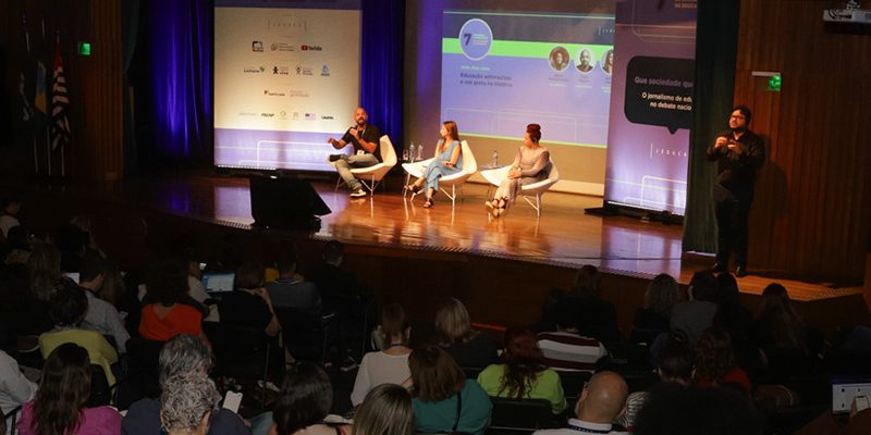 Imagem mostra de forma ampla o auditório e palco em que ocorreu o congresso da Jeduca. No palco, há 3 palestrastes, e ao lado um interprete de libras