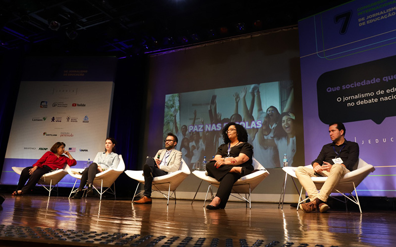 Imagem mostra o palco do congresso da Jeduca com quatro palestrantes, dois homens brancos, duas mulheres brancas e uma mulher negra. Todos estão sentados