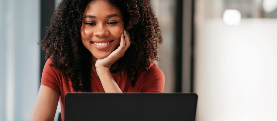 Imagem mostra uma jovem negra, ela tem cabelos compridos, solto, e ela está olhando para um notebook, com a mão esquerda no queixo