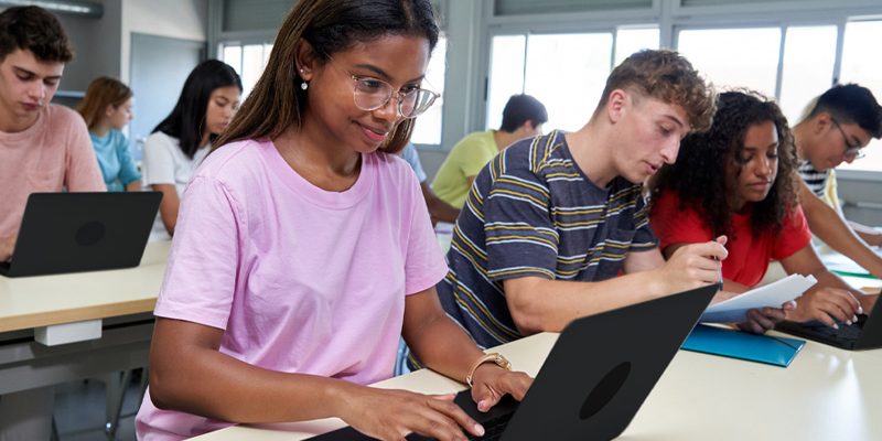 A imagem mostra um grupo diverso de estudantes adolescentes. Eles estão sentados em duas bancadas de estudos com cadernos, canetas e notebooks. Em primeiro plano, a imagem mostra quatro estudantes: duas meninas e dois meninos. Em segundo plano, é possível ver mais quatro estudantes, também dois meninos e duas meninas.