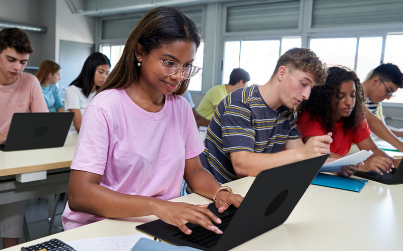 A imagem mostra um grupo diverso de estudantes adolescentes. Eles estão sentados em duas bancadas de estudos com cadernos, canetas e notebooks. Em primeiro plano, a imagem mostra quatro estudantes: duas meninas e dois meninos. Em segundo plano, é possível ver mais quatro estudantes, também dois meninos e duas meninas.