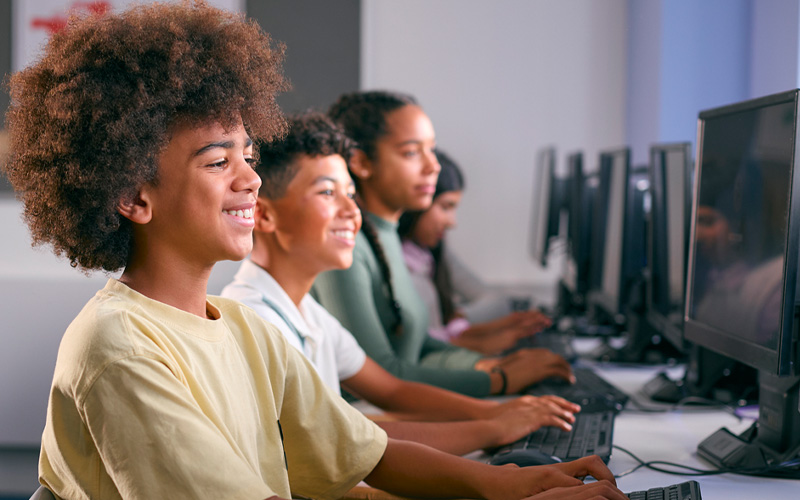 Imagem mostra quatro estudantes em perspectiva, todos estão em sala de aula utilizando computadores