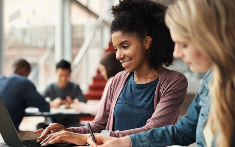A imagem mostra uma estudante negra que veste camiseta azul e blusa vermelha sorrindo e digitando em um notebook preto. Em desfoca, uma estudante loira que veste camisa jeans está com um lápis na mão.